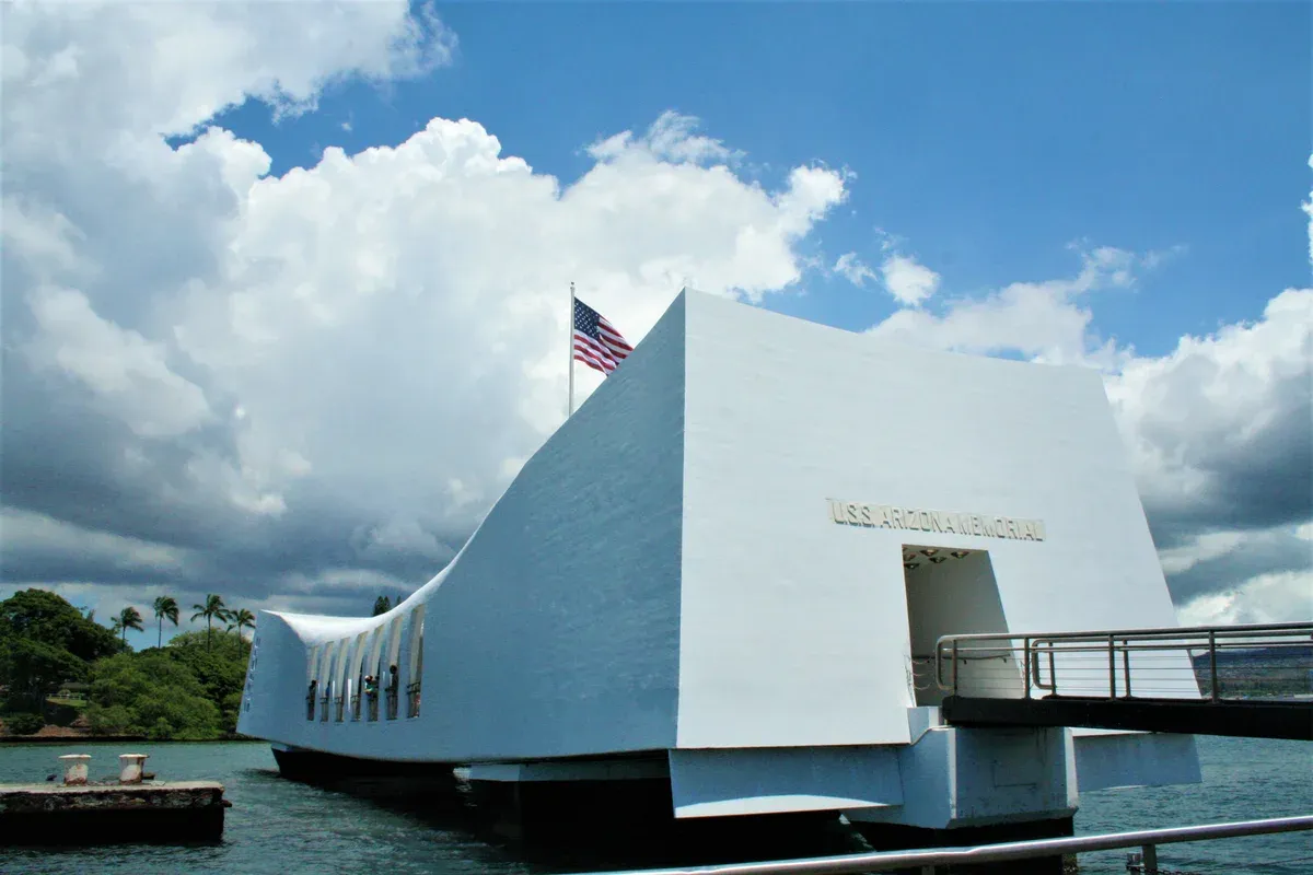 The white USS Arizona Memorial at water level with its entrance, flag and visitor gangway, from the harbor side, a stop on Pearl Harbor tours from Honolulu