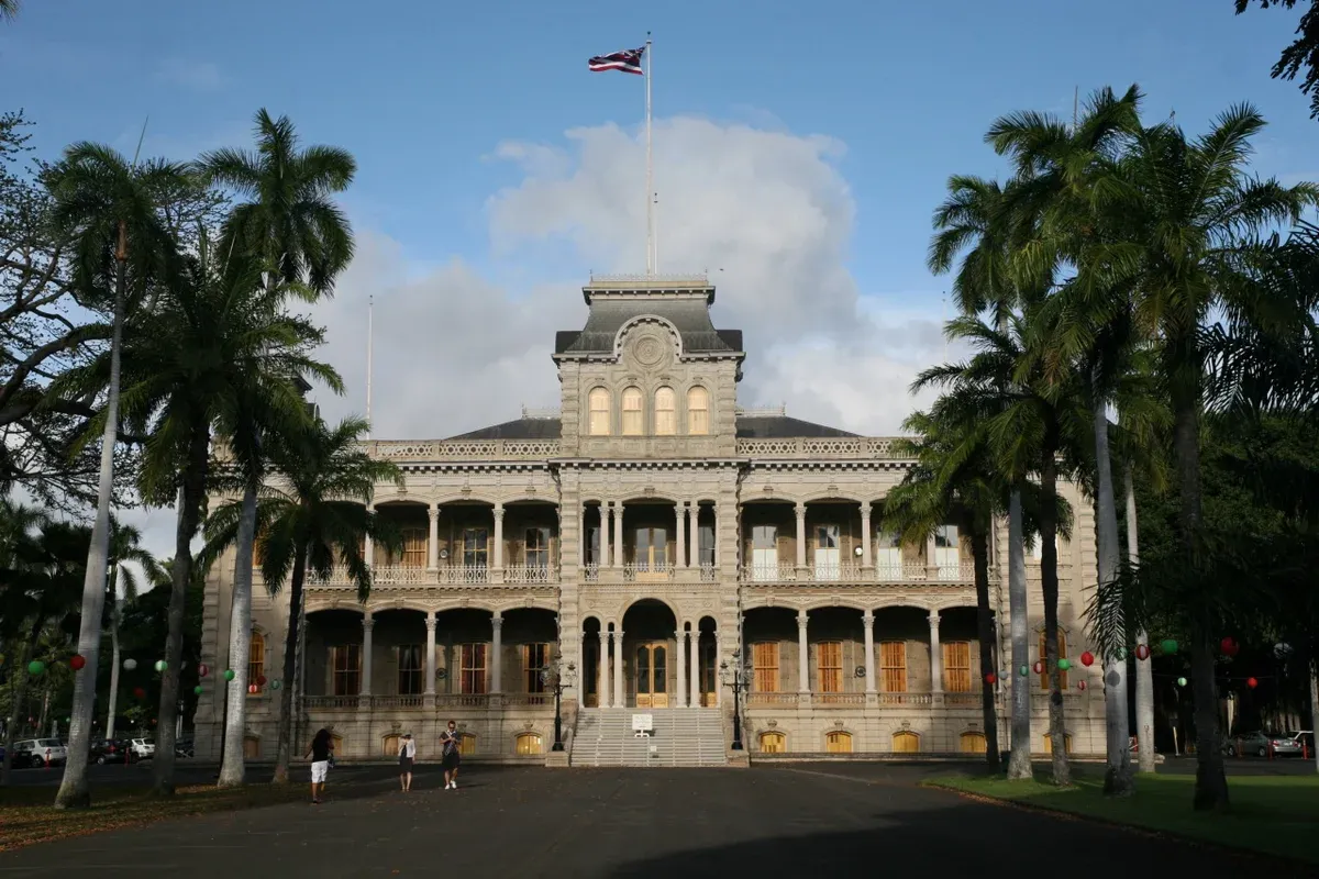 Front facade of Iolani Palace in Honolulu with the Hawaiian flag, royal palms and the forecourt in evening light, a stop on Pearl Harbor tours from Honolulu
